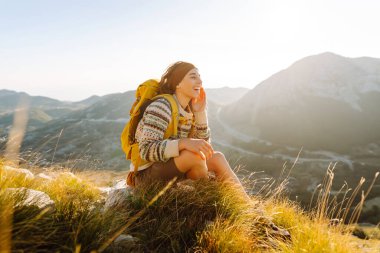 Sarı sırt çantalı güzel bir bayan yürüyüşçü dağ sırtında durup günbatımını izliyor. Genç kadın yolculuğun tadını çıkarıyor ve gün batımının özgürlüğünü hissediyor. Yürüyüş, özgürlük ve aktif yaşam tarzı kavramı.