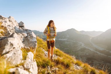 Sarı sırt çantalı güzel bir bayan yürüyüşçü dağ sırtında durup günbatımını izliyor. Genç kadın yolculuğun tadını çıkarıyor ve gün batımının özgürlüğünü hissediyor. Yürüyüş, özgürlük ve aktif yaşam tarzı kavramı.