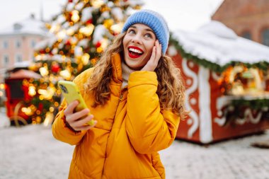 Telefon kullanan bir kadının portresi, pazarda Noel atmosferinin tadını çıkarıyor. Güzel bir kadın yeni yıl pazarında ışıklarla süslenmiş selfie ve blog alıyor. Tatil ve blog konsepti.
