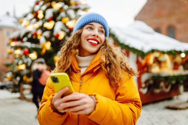 Telefon kullanan bir kadının portresi, pazarda Noel atmosferinin tadını çıkarıyor. Güzel bir kadın yeni yıl pazarında ışıklarla süslenmiş selfie ve blog alıyor. Tatil ve blog konsepti.