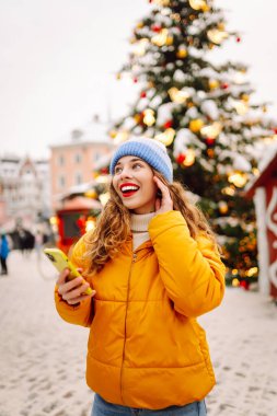 Telefon kullanan bir kadının portresi, pazarda Noel atmosferinin tadını çıkarıyor. Güzel bir kadın yeni yıl pazarında ışıklarla süslenmiş selfie ve blog alıyor. Tatil ve blog konsepti.