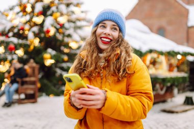 Telefon kullanan bir kadının portresi, pazarda Noel atmosferinin tadını çıkarıyor. Güzel bir kadın yeni yıl pazarında ışıklarla süslenmiş selfie ve blog alıyor. Tatil ve blog konsepti.