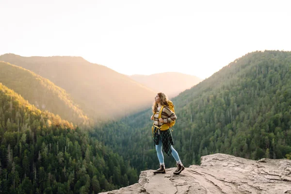 Sarı sırt çantalı genç yürüyüşçü gün batımında uçurumun kenarında duruyor. Gerçek manzara. Güzel bir kadın dağ manzarasından hoşlanır, doğanın özgürlüğünü hisseder. Özgürlük, rahatlama ve macera kavramları..