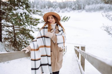 Sıcak bir eşarba sarılmış, şık bir şapka takmış, karlı bir günde tahta bir yolda duran sevimli bir kadın. Dişi bir yürüyüşçü kış manzarasının tadını çıkarır. Macera ve doğa kavramı.