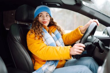 Güneşli kış gününde modern araba kullanan parlak sarı ceketli genç bir kadının portresi. Kadın sürücü, pencerenin dışında karlı bir manzarayla gezmekten hoşlanır. Ulaşım, taksi, soğuk.