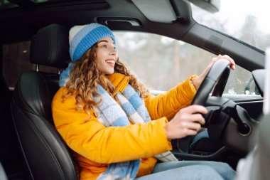 Güneşli kış gününde modern araba kullanan parlak sarı ceketli genç bir kadının portresi. Kadın sürücü, pencerenin dışında karlı bir manzarayla gezmekten hoşlanır. Ulaşım, taksi, soğuk.