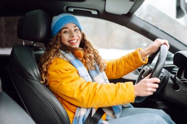 Güneşli kış gününde modern araba kullanan parlak sarı ceketli genç bir kadının portresi. Kadın sürücü, pencerenin dışında karlı bir manzarayla gezmekten hoşlanır. Ulaşım, taksi, soğuk.
