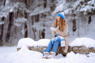 Parlak atkı ve şapkalı gülümseyen bir kadın karlı ormanda termostan sıcak bir içecek dolduruyor. Güzel bir kadın kış manzarasından zevk alır ve çay içer. Doğanın kavramsal görüntüleri, rahatlama, içkiler..