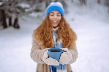 Parlak atkı ve şapkalı gülümseyen bir kadın karlı ormanda termostan sıcak bir içecek dolduruyor. Güzel bir kadın kış manzarasından zevk alır ve çay içer. Doğanın kavramsal görüntüleri, rahatlama, içkiler..