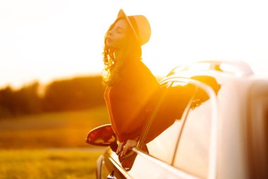 Güneşli bir günde gün batımında neşeli bir kadın arabanın camından sarkıyor. Şapkalı genç bir kadın doğanın tadını çıkarır ve yolculuk özgürlüğünü hisseder. Macera ve rahatlama kavramı.