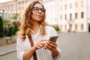 Günbatımında şehrin merkez meydanında telefonu olan şık bir kadının portresi. Güzel bir kadın güneşli bir günde gezintiden hoşlanır. Tarz, moda ve blog kavramı.