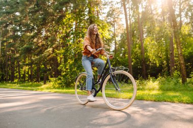 Neşeli bir kadın canlı yeşil bir ormanda bir yol boyunca bisiklet sürüyor. Genç bir kadın güneşli bir günde parkta bisiklet sürmekten hoşlanır. Konsept: boş zaman, hafta sonu. Etkin yaşam biçimi.