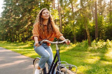Neşeli bir kadın canlı yeşil bir ormanda bir yol boyunca bisiklet sürüyor. Genç bir kadın güneşli bir günde parkta bisiklet sürmekten hoşlanır. Konsept: boş zaman, hafta sonu. Etkin yaşam biçimi.