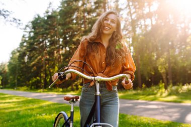 Neşeli bir kadın canlı yeşil bir ormanda bir yol boyunca bisiklet sürüyor. Genç bir kadın güneşli bir günde parkta bisiklet sürmekten hoşlanır. Konsept: boş zaman, hafta sonu. Etkin yaşam biçimi.