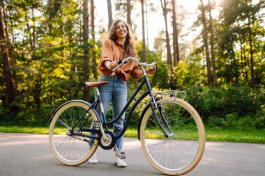 Neşeli bir kadın canlı yeşil bir ormanda bir yol boyunca bisiklet sürüyor. Genç bir kadın güneşli bir günde parkta bisiklet sürmekten hoşlanır. Konsept: boş zaman, hafta sonu. Etkin yaşam biçimi.