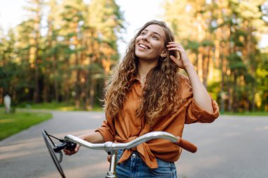 Neşeli bir kadın canlı yeşil bir ormanda bir yol boyunca bisiklet sürüyor. Genç bir kadın güneşli bir günde parkta bisiklet sürmekten hoşlanır. Konsept: boş zaman, hafta sonu. Etkin yaşam biçimi.