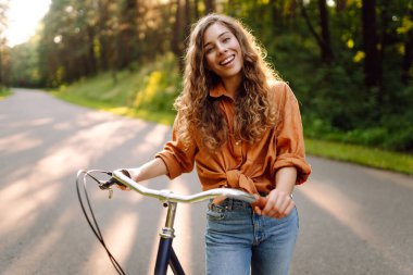 Neşeli bir kadın canlı yeşil bir ormanda bir yol boyunca bisiklet sürüyor. Genç bir kadın güneşli bir günde parkta bisiklet sürmekten hoşlanır. Konsept: boş zaman, hafta sonu. Etkin yaşam biçimi.
