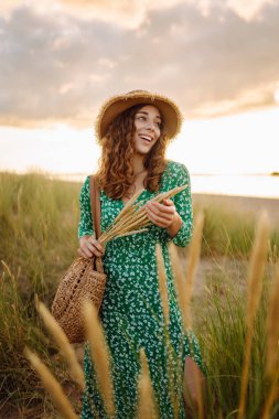 Şık elbiseli ve şapkalı genç bir kadın gün batımında sahilde duruyor. Güzel bir kadın deniz manzarasını sever, dışarıda dinlenir. Konsept: boş zaman, moda.