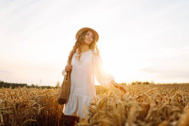 Beyaz elbiseli ve hasır şapkalı genç bir kadın gün batımında altın buğday tarlasında yürüyor. Güzel bir kadın gün batımında bir tarlada poz veriyor. Moda, tarz ve zevk kavramı.