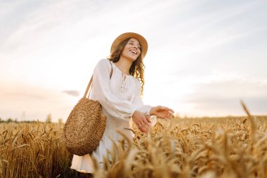 Beyaz elbiseli ve hasır şapkalı genç bir kadın gün batımında altın buğday tarlasında yürüyor. Güzel bir kadın gün batımında bir tarlada poz veriyor. Moda, tarz ve zevk kavramı.