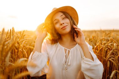 Beyaz elbiseli ve hasır şapkalı genç bir kadın gün batımında altın buğday tarlasında yürüyor. Güzel bir kadın gün batımında bir tarlada poz veriyor. Moda, tarz ve zevk kavramı.