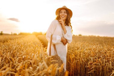 Beyaz elbiseli ve hasır şapkalı genç bir kadın gün batımında altın buğday tarlasında yürüyor. Güzel bir kadın gün batımında bir tarlada poz veriyor. Moda, tarz ve zevk kavramı.
