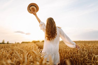 Beyaz elbiseli ve hasır şapkalı genç bir kadın gün batımında altın buğday tarlasında yürüyor. Güzel bir kadın gün batımında bir tarlada poz veriyor. Moda, tarz ve zevk kavramı.