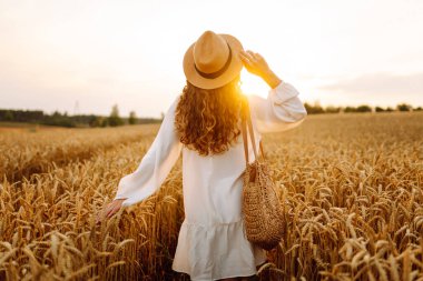 Beyaz elbiseli ve hasır şapkalı genç bir kadın gün batımında altın buğday tarlasında yürüyor. Güzel bir kadın gün batımında bir tarlada poz veriyor. Moda, tarz ve zevk kavramı.