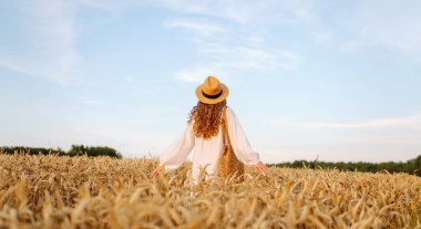 Beyaz elbiseli ve hasır şapkalı genç bir kadın gün batımında altın buğday tarlasında yürüyor. Güzel bir kadın gün batımında bir tarlada poz veriyor. Moda, tarz ve zevk kavramı.