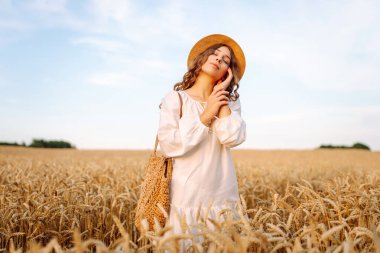 Beyaz elbiseli ve hasır şapkalı genç bir kadın gün batımında altın buğday tarlasında yürüyor. Güzel bir kadın gün batımında bir tarlada poz veriyor. Moda, tarz ve zevk kavramı.