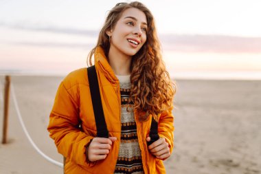 Parlak sarı ceketli gülümseyen bir kadın gün batımında kumlu bir plajda geziniyor. Sırt çantalı bir bayan yürüyüşçü deniz manzarasının tadını çıkarıyor. Yürüyüş ve seyahat kavramı. Yaşam biçimi.