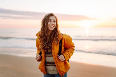 Parlak sarı ceketli gülümseyen bir kadın gün batımında kumlu bir plajda geziniyor. Sırt çantalı bir bayan yürüyüşçü deniz manzarasının tadını çıkarıyor. Yürüyüş ve seyahat kavramı. Yaşam biçimi.