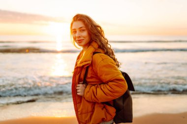 Parlak sarı ceketli gülümseyen bir kadın gün batımında kumlu bir plajda geziniyor. Sırt çantalı bir bayan yürüyüşçü deniz manzarasının tadını çıkarıyor. Yürüyüş ve seyahat kavramı. Yaşam biçimi.