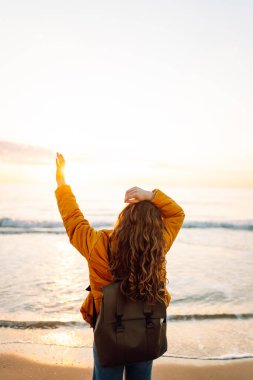 Parlak sarı ceketli gülümseyen bir kadın gün batımında kumlu bir plajda geziniyor. Sırt çantalı bir bayan yürüyüşçü deniz manzarasının tadını çıkarıyor. Yürüyüş ve seyahat kavramı. Yaşam biçimi.