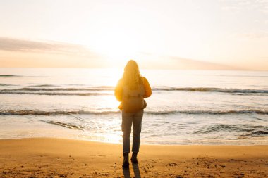 Parlak sarı ceketli gülümseyen bir kadın gün batımında kumlu bir plajda geziniyor. Sırt çantalı bir bayan yürüyüşçü deniz manzarasının tadını çıkarıyor. Yürüyüş ve seyahat kavramı. Yaşam biçimi.