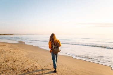 Parlak sarı ceketli gülümseyen bir kadın gün batımında kumlu bir plajda geziniyor. Sırt çantalı bir bayan yürüyüşçü deniz manzarasının tadını çıkarıyor. Yürüyüş ve seyahat kavramı. Yaşam biçimi.