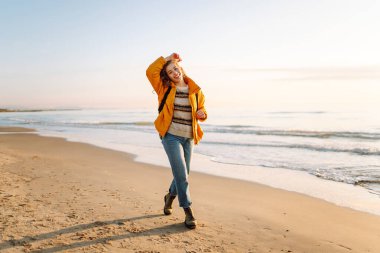 Parlak sarı ceketli gülümseyen bir kadın gün batımında kumlu bir plajda geziniyor. Sırt çantalı bir bayan yürüyüşçü deniz manzarasının tadını çıkarıyor. Yürüyüş ve seyahat kavramı. Yaşam biçimi.