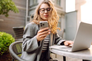 Gözlüklü ve ceketli genç bir kadın, elinde telefon, masada dizüstü bilgisayarla oturuyor. Tarz sahibi bayan serbest yazar terasta telefonla ve dizüstü bilgisayarla çalışmaktan zevk alır. Uzak çalışma kavramı, günlük tutma