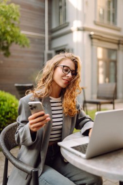Gözlüklü ve ceketli genç bir kadın, elinde telefon, masada dizüstü bilgisayarla oturuyor. Tarz sahibi bayan serbest yazar terasta telefonla ve dizüstü bilgisayarla çalışmaktan zevk alır. Uzak çalışma kavramı, günlük tutma