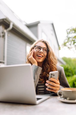 Gözlüklü ve ceketli genç bir kadın, elinde telefon, masada dizüstü bilgisayarla oturuyor. Tarz sahibi bayan serbest yazar terasta telefonla ve dizüstü bilgisayarla çalışmaktan zevk alır. Uzak çalışma kavramı, günlük tutma