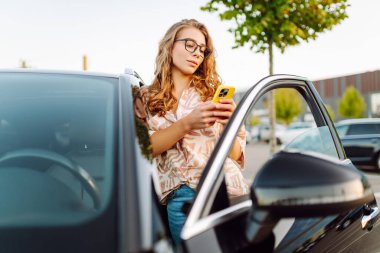 Neşeli bir kadın güneşli bir günde siyah bir arabanın yanında akıllı telefonuna bakıyor. Akıllı telefonlu genç bir bayan sürücü bir otoparkta dinleniyor. Kavram: teknoloji, kentsel.