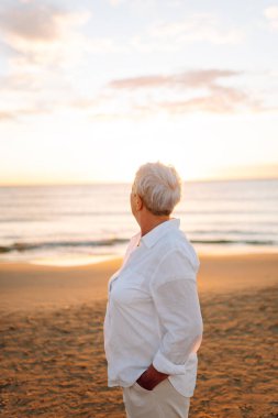 Neşeli yaşlı bir kadın gün batımında kumlu bir plajda dolaşırken kendini özgür hisseder. Gri saçlı güzel bir kadın deniz kıyısında yalınayak yürümekten hoşlanıyor. Doğa, yaşlanma, emeklilik kavramları.