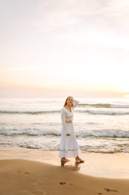 Beyaz elbiseli mutlu bir kadın gün batımında sahilde geziniyor. Genç kadın deniz manzarasının tadını çıkarıyor ve açık havanın özgürlüğünü hissediyor. Doğa kavramı, keyif.