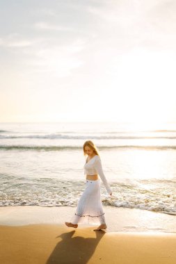 Beyaz elbiseli mutlu bir kadın gün batımında sahilde geziniyor. Genç kadın deniz manzarasının tadını çıkarıyor ve açık havanın özgürlüğünü hissediyor. Doğa kavramı, keyif.
