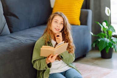 Elinde kitap olan genç bir kadının portresi, rahat bir odada kanepede dinleniyor. Gülümseyen bir kadın güneşli bir günde evde kitap okumaktan hoşlanır. Konseptler: eğitim, rahatlama.