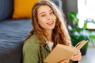 Elinde kitap olan genç bir kadının portresi, rahat bir odada kanepede dinleniyor. Gülümseyen bir kadın güneşli bir günde evde kitap okumaktan hoşlanır. Konseptler: eğitim, rahatlama.
