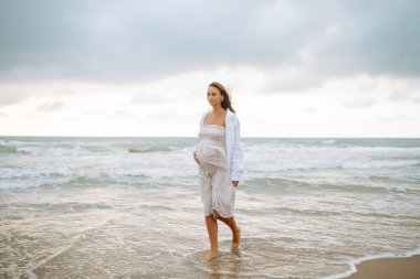 Sevimli bir elbise içinde neşeli bir anne gün batımında kumsalda geziniyor. Hamile kadın deniz manzarasından zevk alır ve özgür hisseder. Fikir: yürüme, rahatlama ve beklenti. Yaşam biçimi.