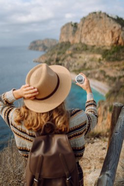 Şapkalı genç bir kadının deniz manzarasına karşı pusula tutuşu. Elinde pusula olan bir kadın uçurumun kenarında duruyor, yol tarifi arıyor. Yürüyüş, yön bulma ve doğa kavramları. Etkin yaşam biçimi.