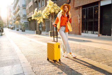 Şapka takan ve sarı bir bavul taşıyan bir turist gün batımında şehir sokaklarında geziniyor. Genç bir kadın bavulla seyahat etmekten hoşlanır. Konseptler: turizm, tatil, hafta sonu.