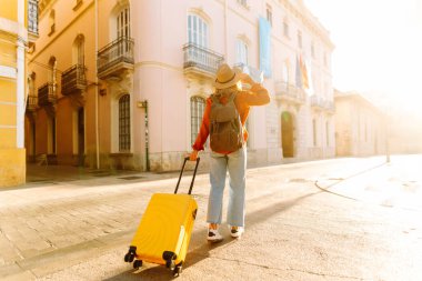 Şapka takan ve sarı bir bavul taşıyan bir turist gün batımında şehir sokaklarında geziniyor. Genç bir kadın bavulla seyahat etmekten hoşlanır. Konseptler: turizm, tatil, hafta sonu.